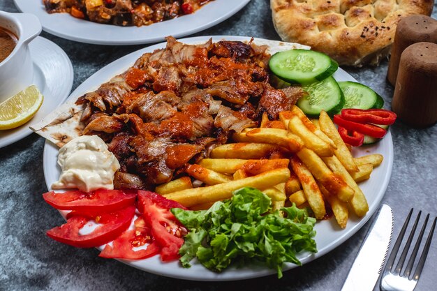 Side view doner on a plate with french fries tomato fresh cucumber yogurt and bread on the table