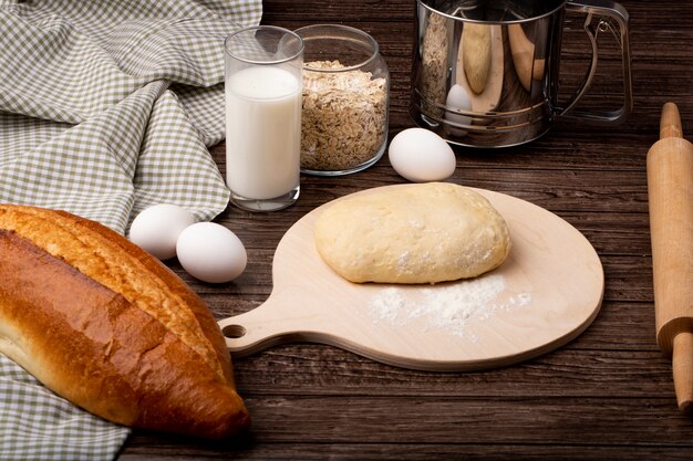 Side view of dough and flour on cutting board with milk eggs baguette oat-flakes on wooden background