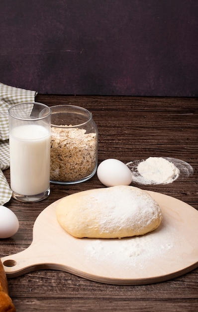 Side view of dough and flour on cutting board with milk oat-flakes and egg on wooden surface and maroon background with copy space