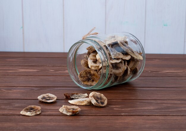 Side view of dried banana chips scattered from a glass jar on wooden background