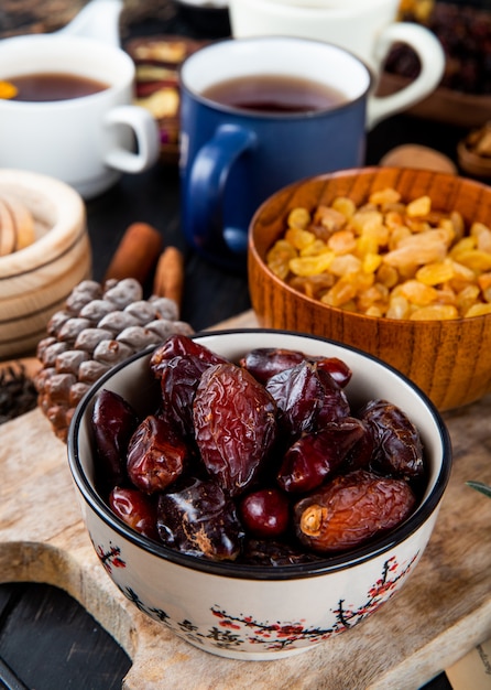 Side view of dried dates in a bowl and yellow raisins in a wood bowl with a mug of tea on the table