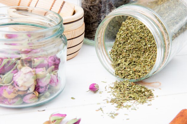 Side view of dried peppermint scattered from a glass jar and rose buds in glass jar on white wood