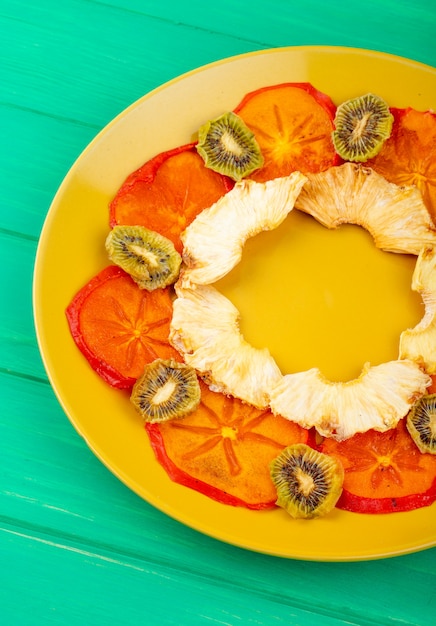 Side view of dried persimmon slices with pineapple and kiwi slices arranged on a yellow plate on green wooden background