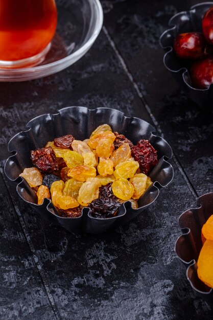 Side view of dried raisins and dried dates in mini tart tins served with tea on black background