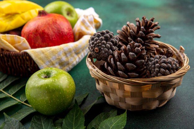 Side view of dry pine cones in a bucket with fresh fruits such as pomegranate apple mango on yellow checked busket with leaf on black surface