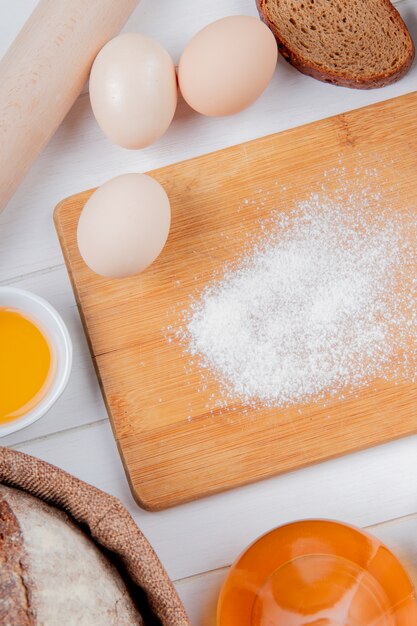 Side view of egg and flour on cutting board with rye and cob bread butter rolling pin on wooden surface