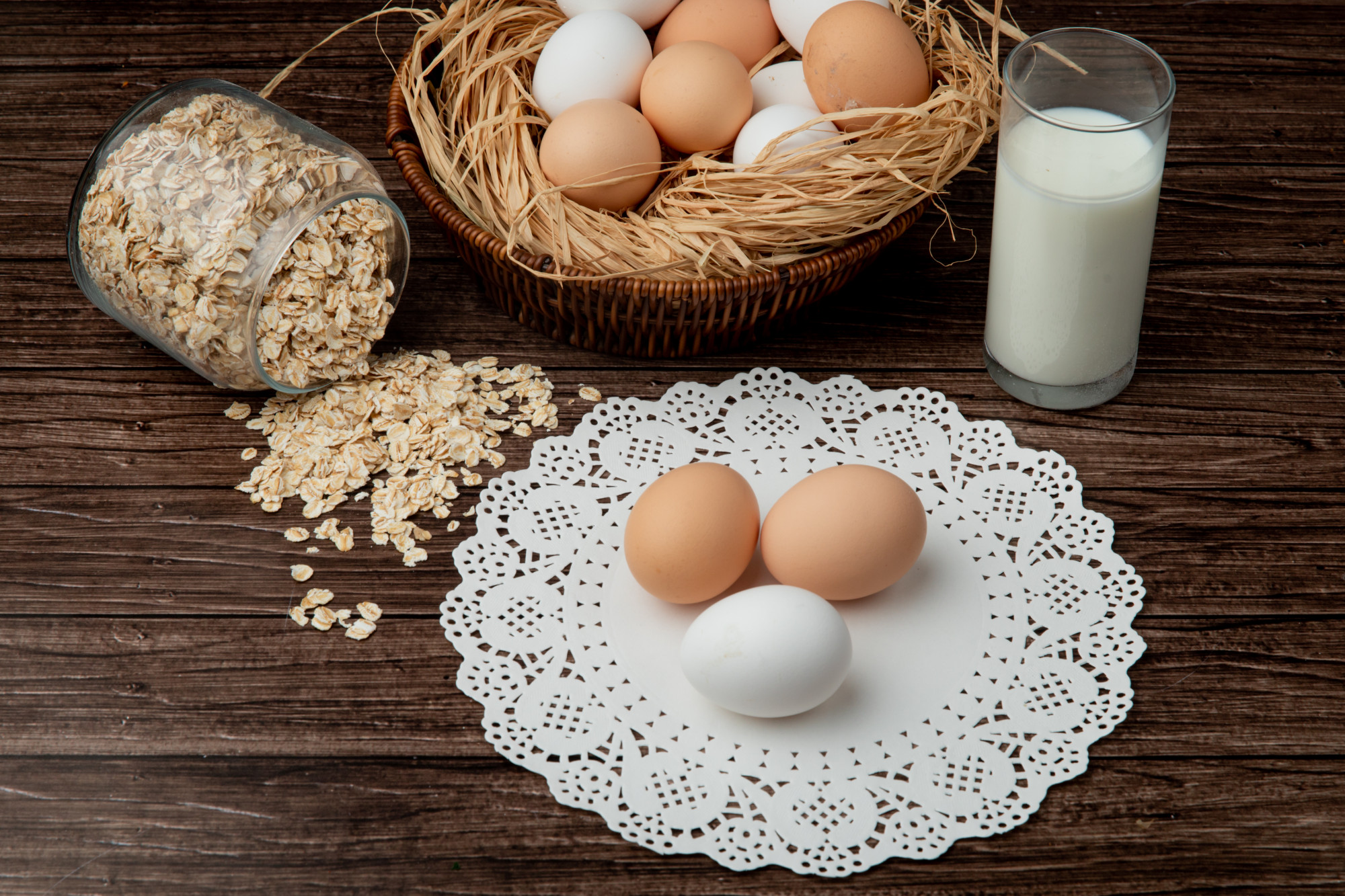 Side view of eggs on paper doily with oat flakes spilling out of jar and milk on wooden background