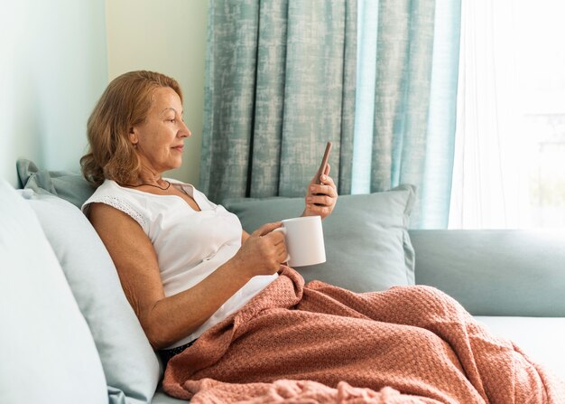Side view of elder woman at home during the pandemic enjoying a cup of coffee and using smartphone