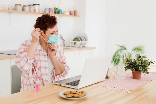 Vista laterale della donna più anziana che indossa la maschera medica a casa prima di lavorare al computer portatile