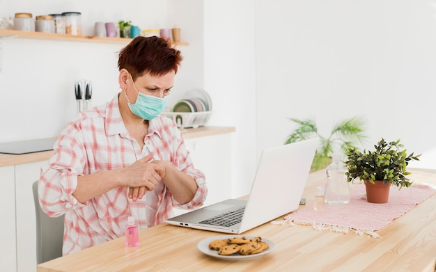 Side view of elder woman with medical mask using hand sanitizer before working on laptop