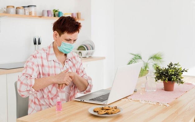Вид сбоку старшей женщины с медицинской маской, используя дезинфицирующее средство для рук, прежде чем работать на ноутбуке