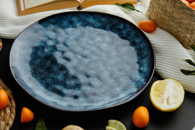 Free photo side view of empty plate with open book and lemon half lime slices kumquats cloth on black background