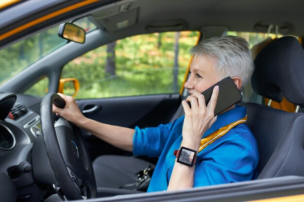 Side view of fashionable mature careless European female with blonde pixie hair sitting in driver's seat with one hand on steering wheel, holding cell phone, talking, concentrating on road