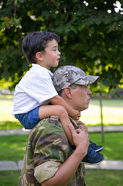 Side view of father holding son on neck and walking in city park. Caucasian son sitting on neck of dad in army uniform, hugging him and looking forward. Fatherhood and returning home concept