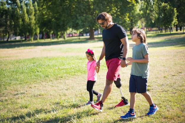 子供と一緒に歩いている障害を持つ父親の側面図。小さな男の子と女の子と一緒に公園を散歩している機械的な脚を持つ波状の髪の男。障害、家族、愛の概念
