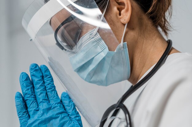 Вид сбоку на женщину-врача с защитной маской и медицинской маской, молящейся