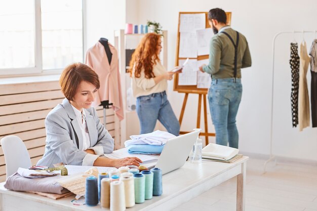 Side view of female fashion designer working in atelier with colleagues and laptop