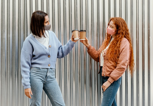 Side view of female friends with face masks outdoors cheering with coffee cups