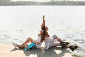 Foto gratuita vista laterale di amiche con skateboard e pattini a rotelle in riva al lago con spazio di copia