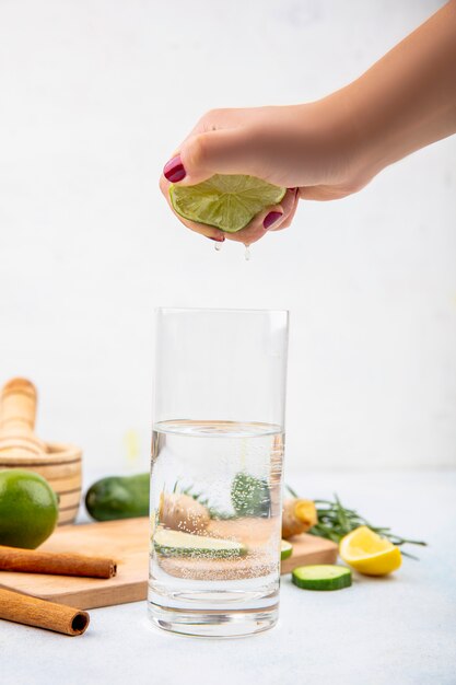 Side view of female hand squeezing fresh lemon into a glass of water on white surface