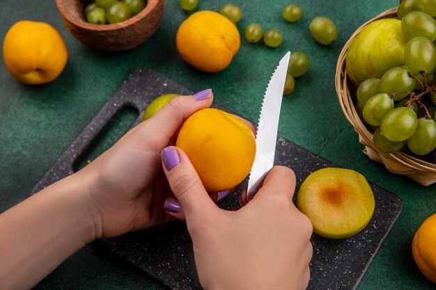 ナイフでネクタコットを切る女性の手の側面図とまな板の半分カットプルオットと緑の背景のボウルにブドウの果実とバスケットのブドウプルオット