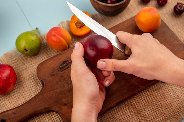 Side view of female hands cutting peach with knife and apricot on cutting board and cherries in bowl peach pear apricot on sackcloth and blue background