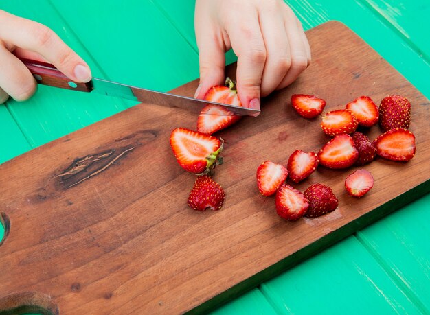 Side view of female hands cutting strawberries with knife on cutting board on green surface