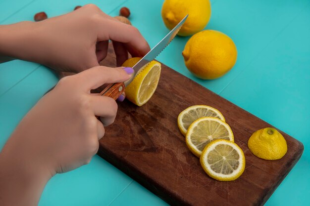 Side view of female hands slicing lemon with knife on cutting board and nuts with lemons on blue background