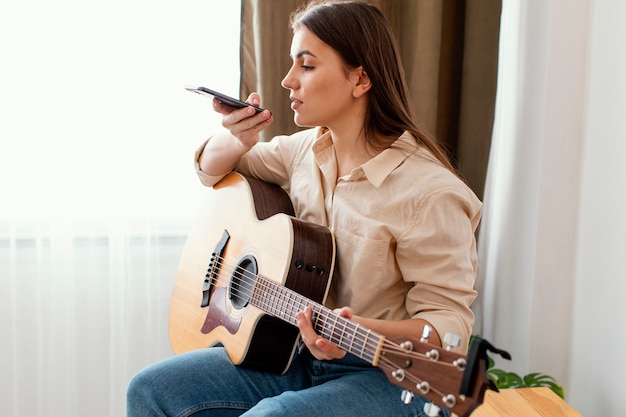 어쿠스틱 기타를 들고 집에서 스마트 폰으로 말하는 여성 음악가의 측면보기