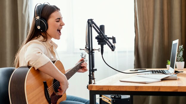 Вид сбоку женского музыканта, записывающего песню дома во время игры на акустической гитаре