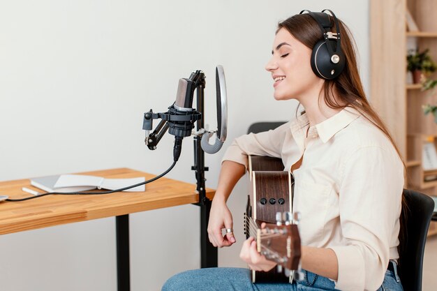 집에서 어쿠스틱 기타를 연주하는 동안 여성 음악가 녹음 노래의 측면보기
