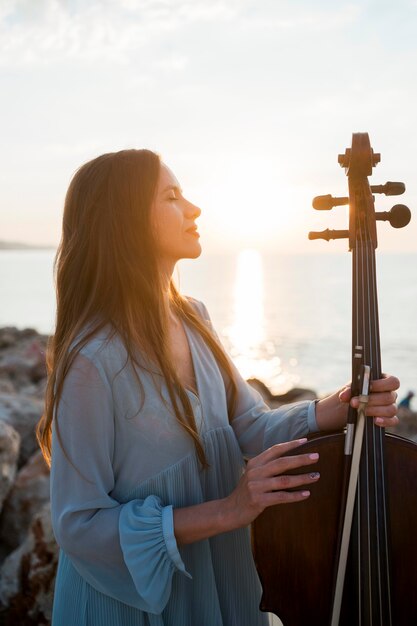 Вид сбоку девушки-музыканта с виолончелью на открытом воздухе на закате