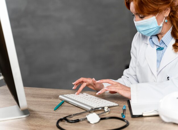 彼女の机にキーボードで書いている医療マスクを持つ女性医師の側面図