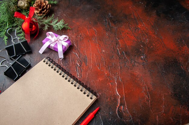 Side view of fir branches decoration accessories and gift next to notebook with pen on dark background