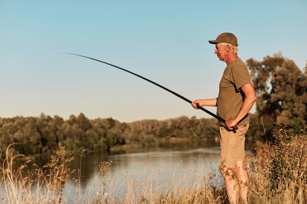 湖や川のほとりに立って、手に釣り竿を見て、日没で、美しい自然の中で、緑のTシャツとズボンを着て釣りをしている漁師の側面図。