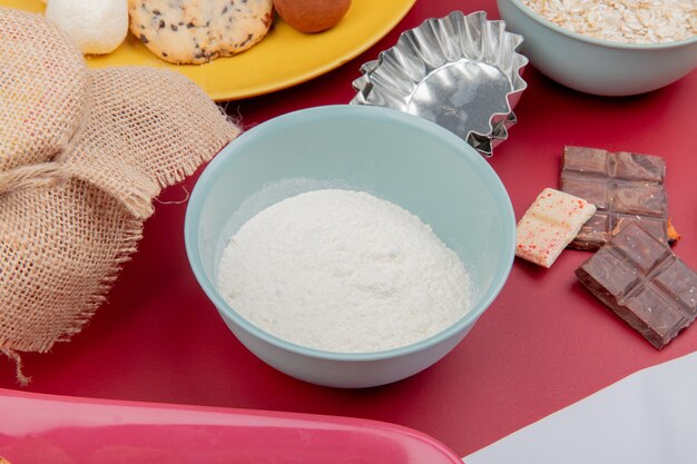 Side view of flour in bowl and chocolates with cookies and oat-flakes on red surface