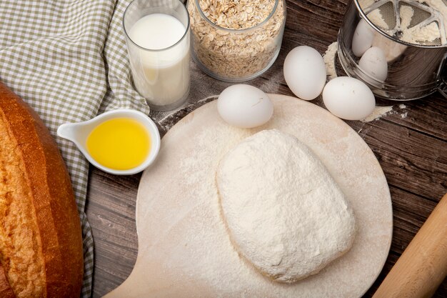 Side view of foods as melted butter milk bread eggs with oat-flakes and dough sprinkled with flour on cutting board on wooden background