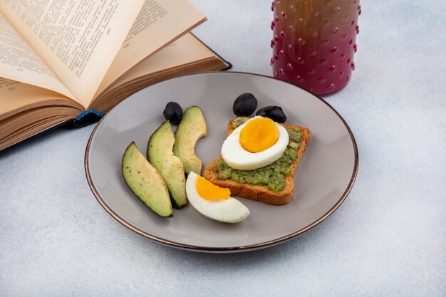 Side view of fresh avocado slices with avocado on a bread slice with poached egg on a plate with cocktail in a glass jar and book on white surface