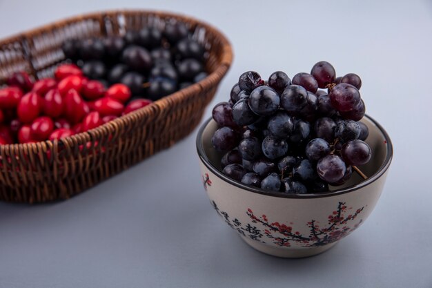 Вид сбоку свежего темнокожего винограда на ведре с ягодами кизила на сером фоне