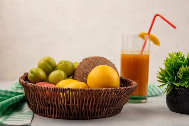 Side view of fresh fruits like green cherry plumscoconutyellow peaches on a bucket with fresh peach juice on a glass on a white background