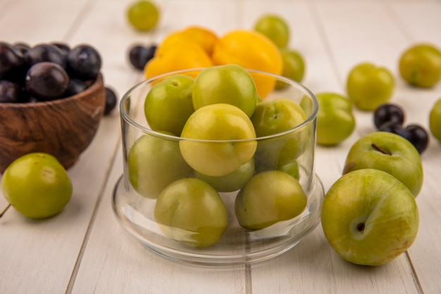 Side view of fresh green cherry plums on a glass bowl with dark purple sloes on a wooden bowl with peaches isolated on a white wooden background