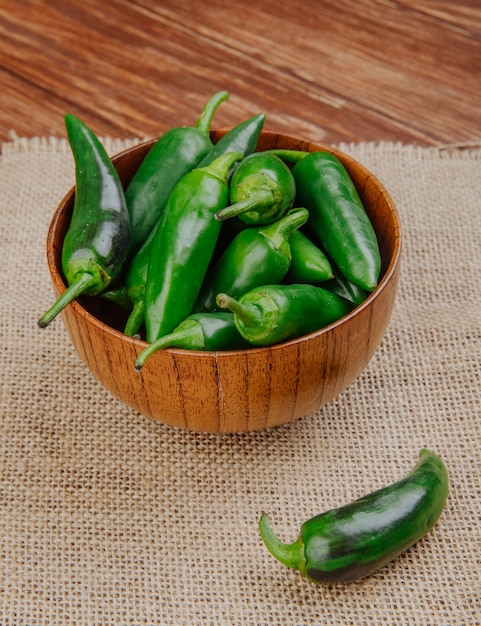 side view of fresh green chili peppers in a wooden bowl on sackcloth on rustic surface