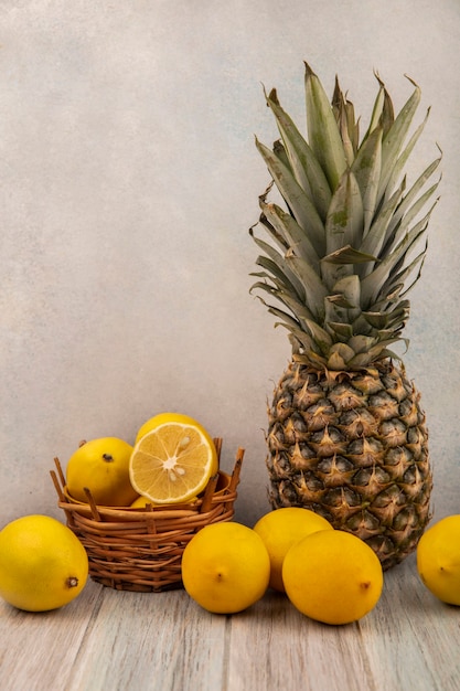 白い表面の灰色の木製のテーブルに分離されたレモンとパイナップルとバケツの上の新鮮なレモンの側面図