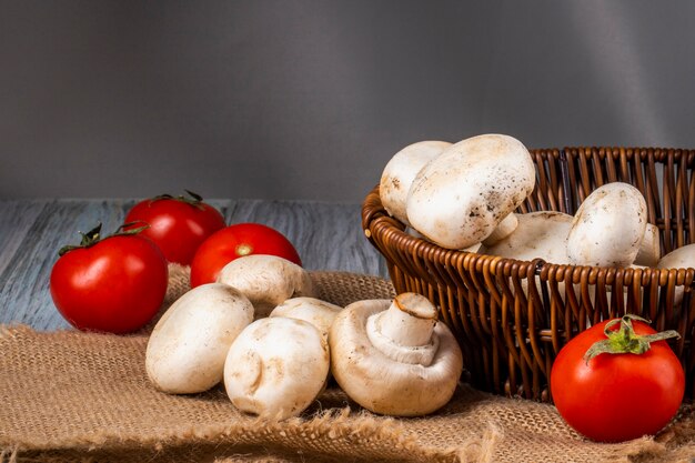 Side view of fresh mushrooms champignon in a wicker basket and fresh tomatoes on sackcloth