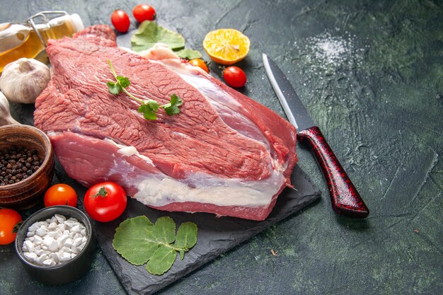 Side view of fresh raw red meat on black tray pepper vegetables fallen oil bottle knife on dark color background