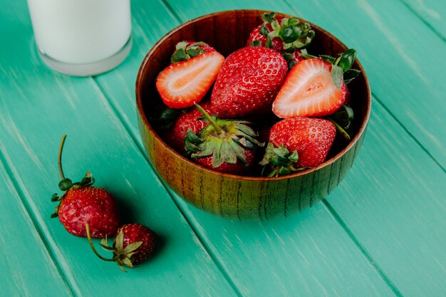 Side view of fresh ripe strawberries in a wood bowl with a glass of milk on green wood