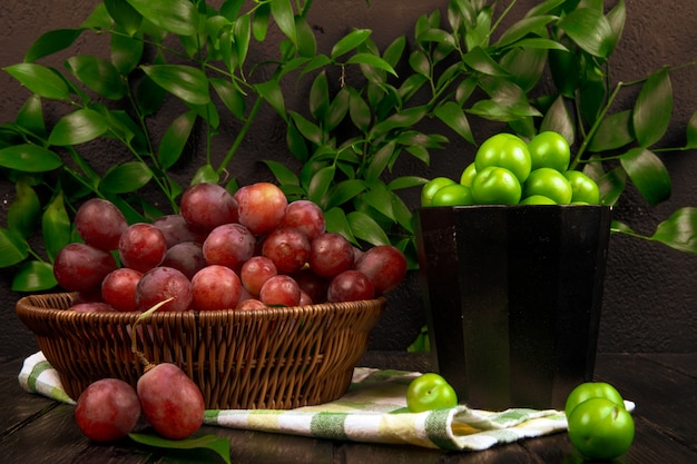 Side view of fresh sweet grape in a wicker basket and a bowl with sour green plums on wooden surface on green leaves table