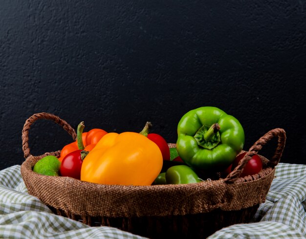 Side view of fresh vegetables colorful bell peppers tomatoes and cucumbers in a wicker basket on plaid fabric on black