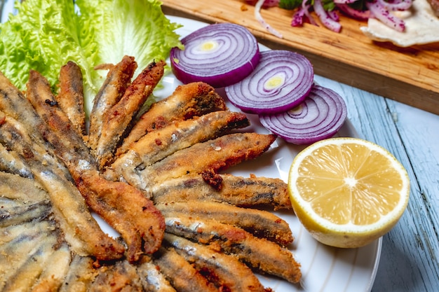 Side view fried anchovy with red onion lettuce and slice of lemon on a plate