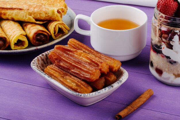 Side view of fried dough pastry with honey served with a cup of green tea and cinnamon sticks on purple wooden table